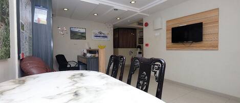 A dining area with a marble table, four chairs, a television mounted on the wall, and a kitchen in the background.