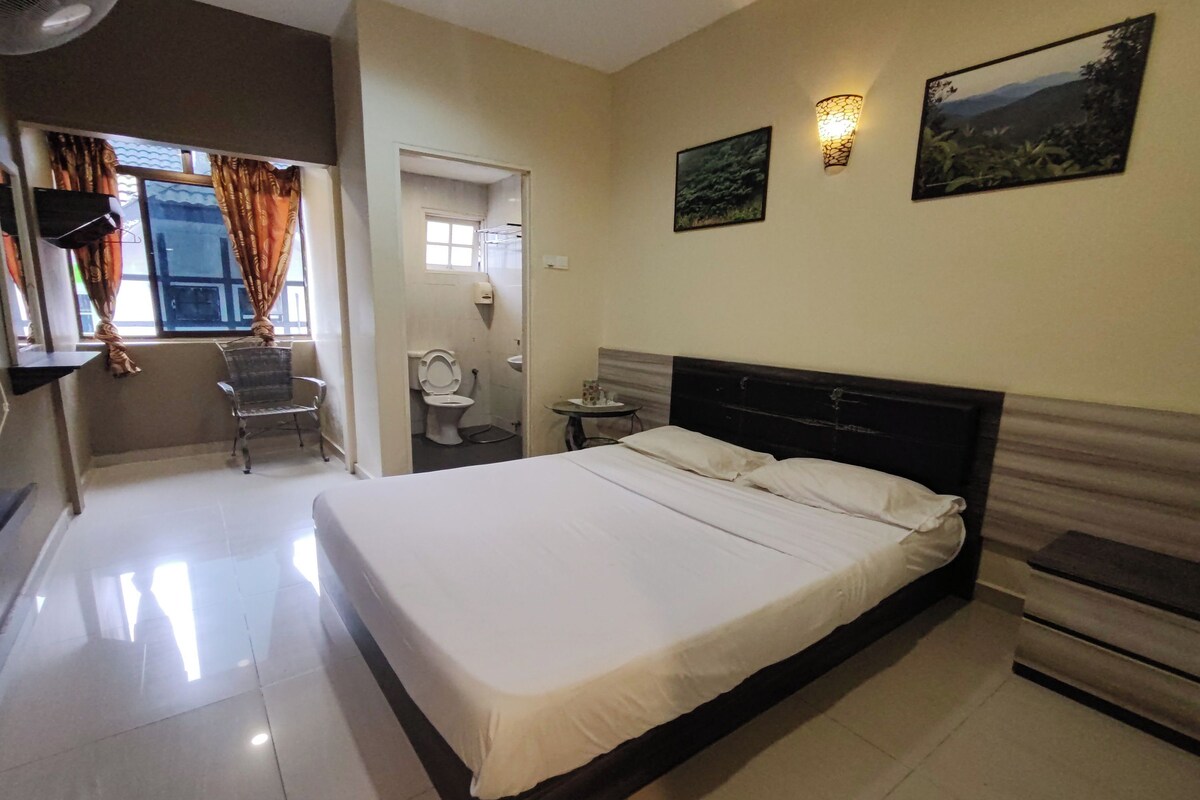 A bedroom with a large bed, a bedside table, a chair, a small table with a lamp, and a window with curtains.