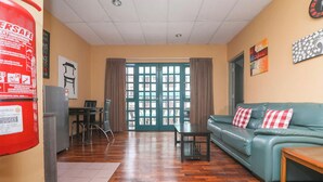 Ein möbliertes Wohnzimmer mit einer Couch, einem Tisch und einem Sessel. An der Wand hängt ein Feuerlöscher, ein Glastür und ein Fenster mit Vorhängen.