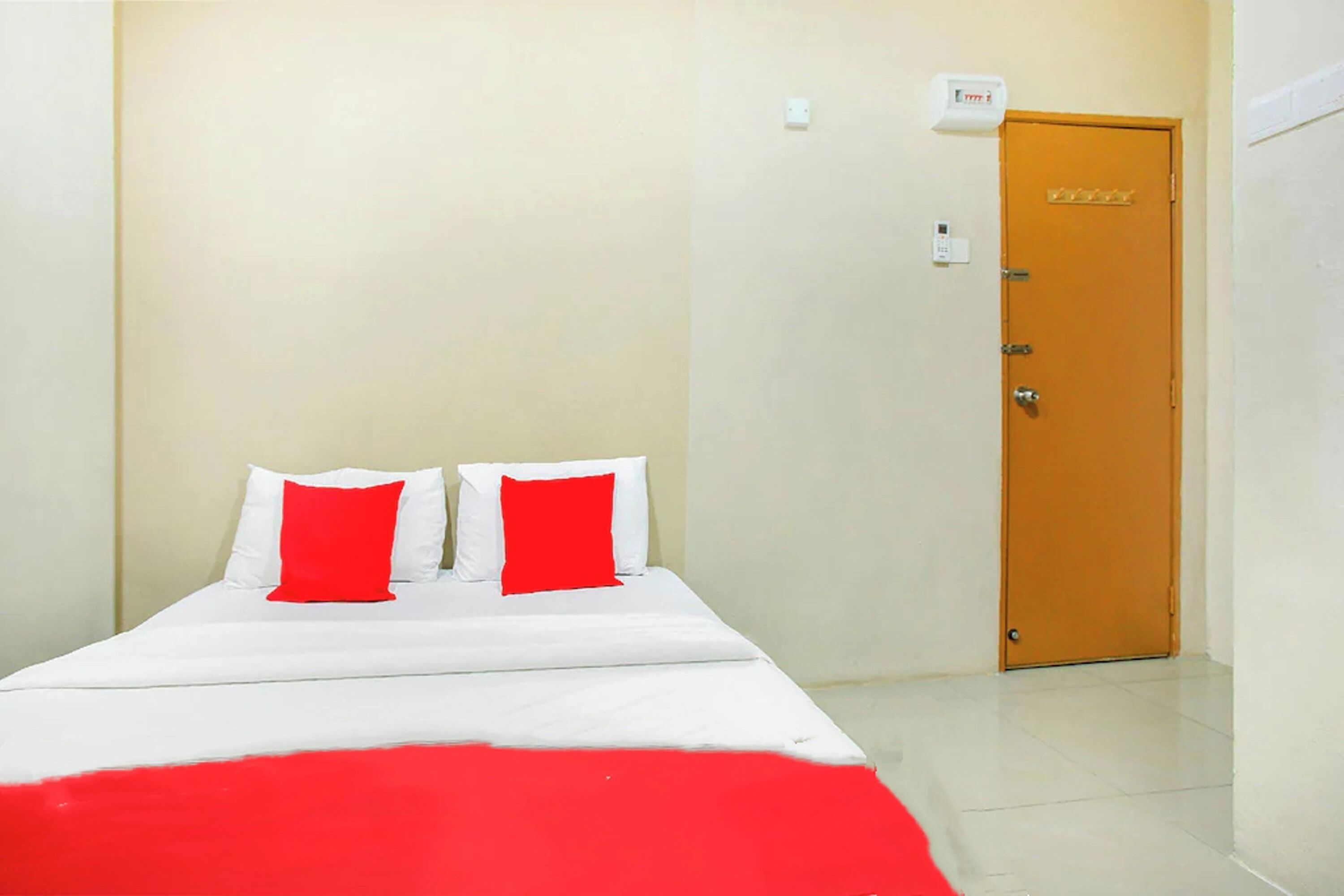 A single bed with white bedding and red pillows, a wooden door, and a light switch on the wall.
