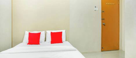 A single bed with white bedding and red pillows, a wooden door, and a light switch on the wall.