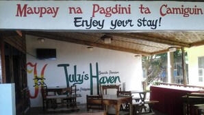 Een terras met houten tafels en stoelen, een bord met de tekst 'Enjoy your stay!' en 'Maupay na Pagdini ta Camiguin', en een muurschildering met de tekst 'Tuly's Haven'.