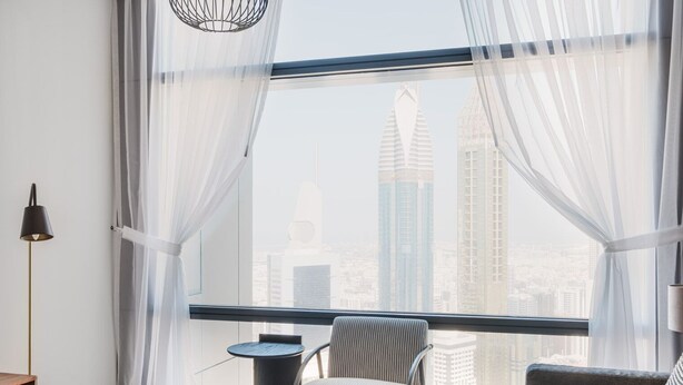 A modern room with a large window, a round table, a chair, and a sofa. - Blueground | DIFC, pool, nr Dubai Mall (Dubai)