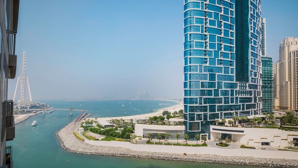 Een modern hotel met een gebogen gevel, gelegen nabij een waterkant met uitzicht op zee en een brug op de achtergrond.
