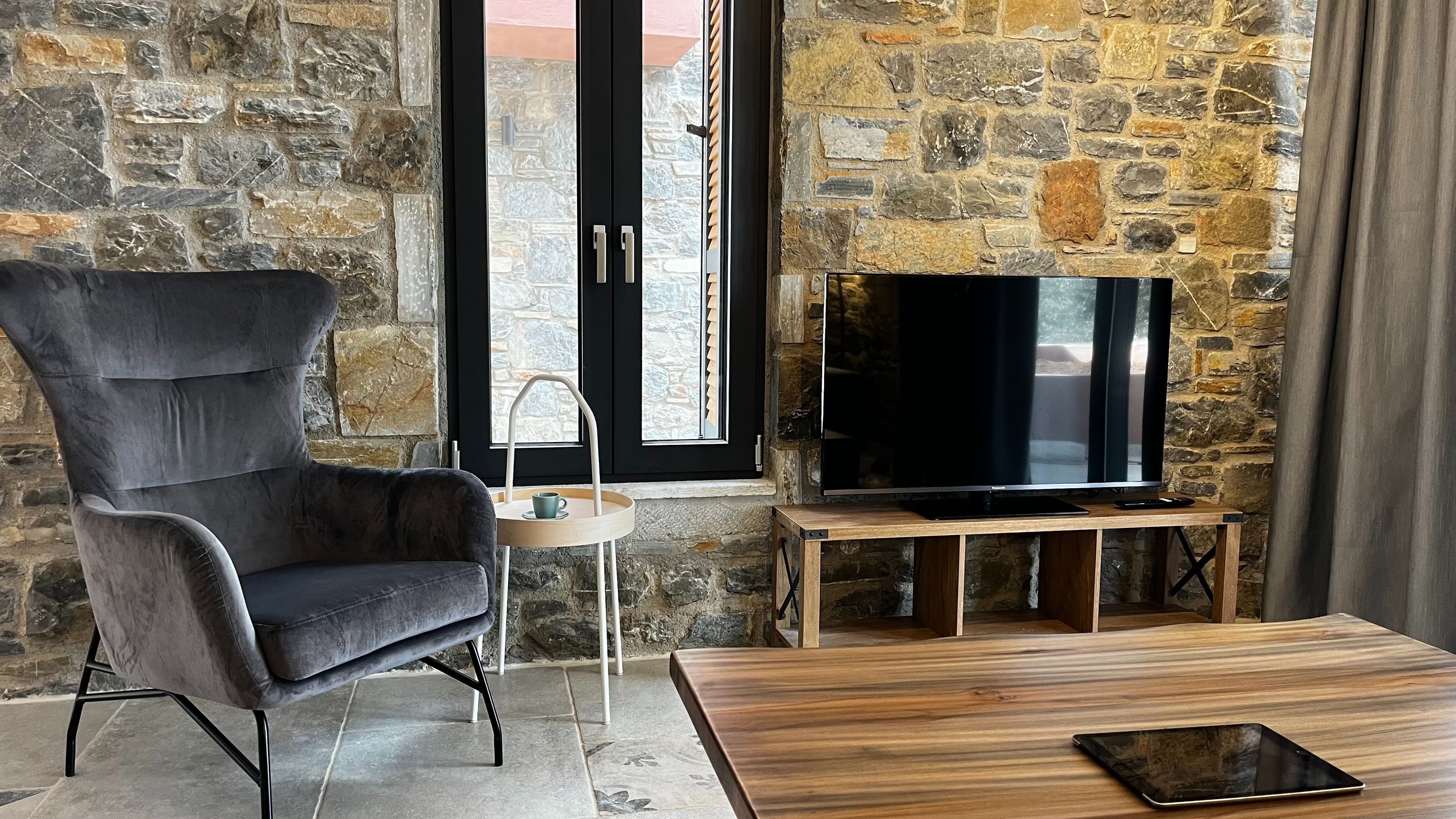 A cozy room with a stone wall, a comfortable armchair, a flat-screen TV on a wooden stand, and a tablet on a coffee table.