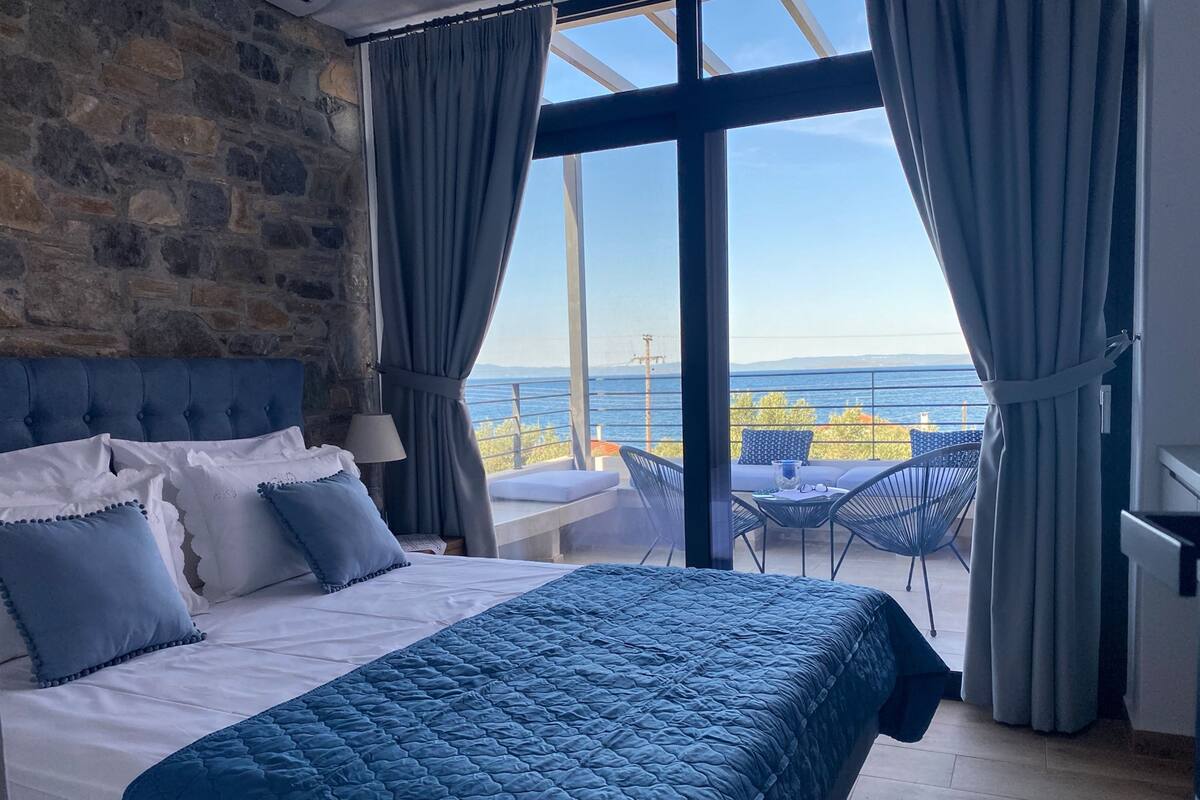 A bedroom with a large bed, a stone wall, and a balcony with a view of the sea.