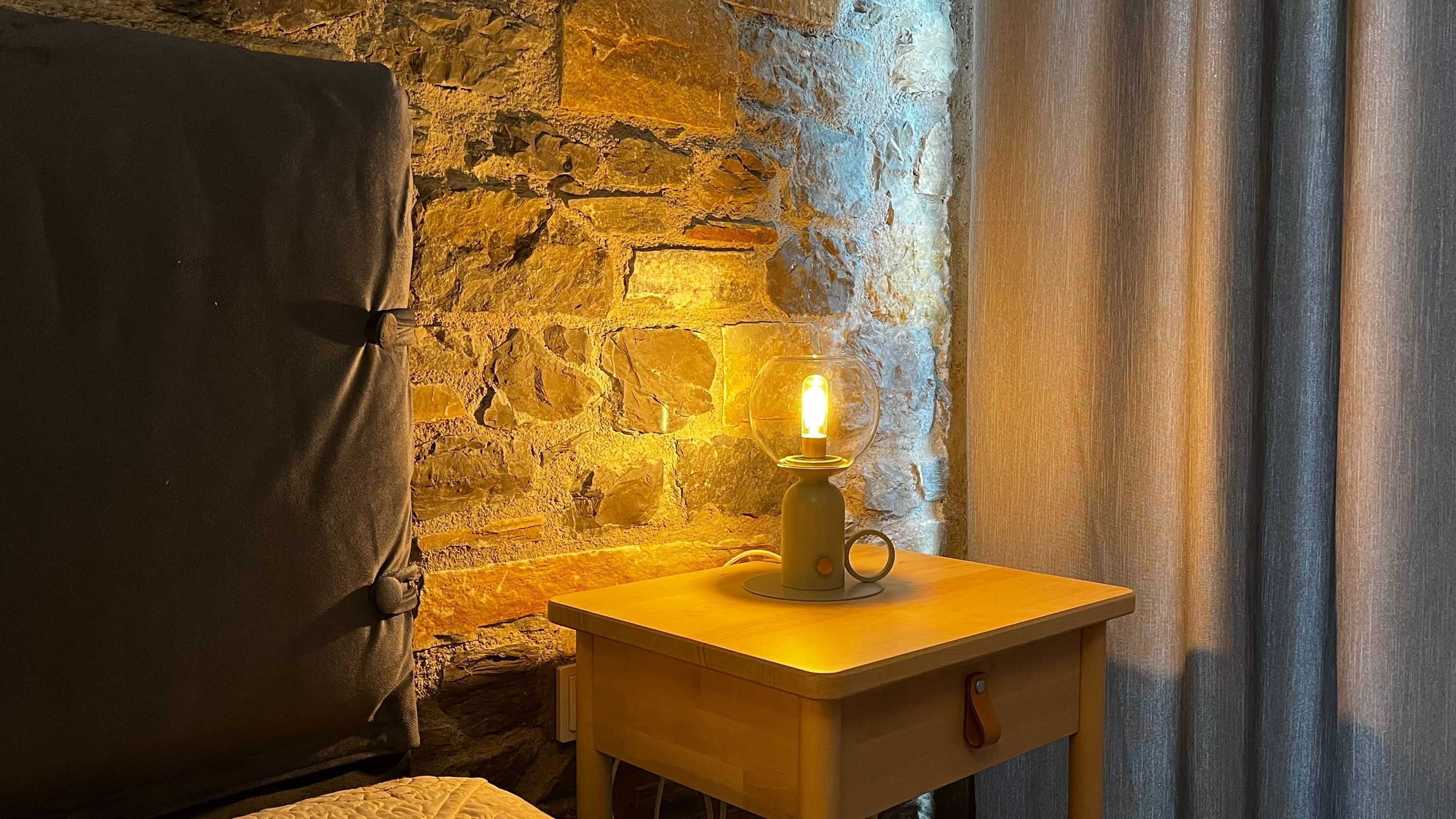 A room with a stone wall, a wooden nightstand with a lit candle, a bed with a dark cover, and a window with curtains.