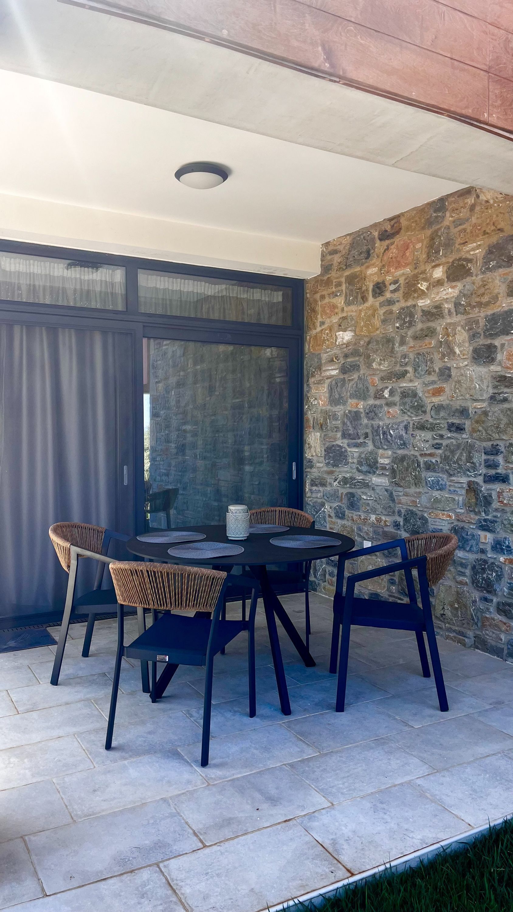 An outdoor patio with a round table and four chairs, a stone wall, and a large window with curtains.