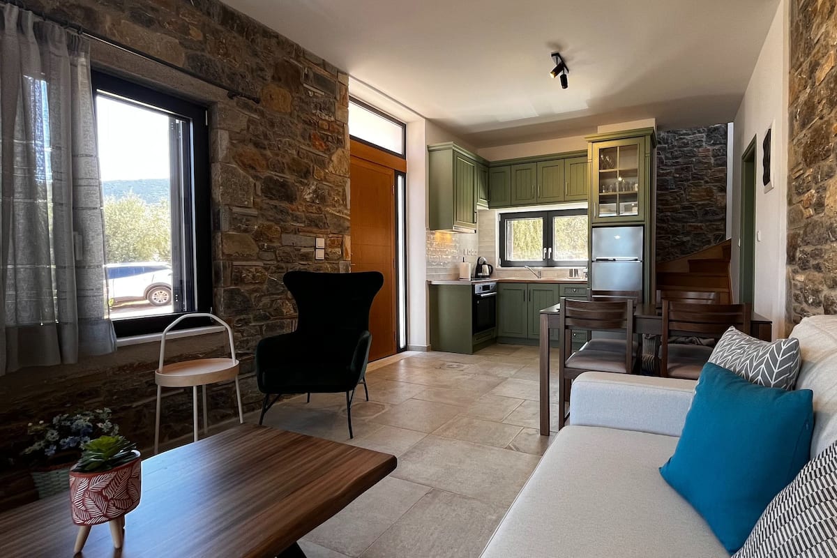 A living room with a stone wall, a wooden dining table, a sofa with patterned cushions, and a kitchen area with green cabinets and a window.