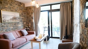 A cozy living room with a stone wall, a sofa, a coffee table with candles, and a view through a glass door. - Aélia Stone Houses in Nies (Almyros)