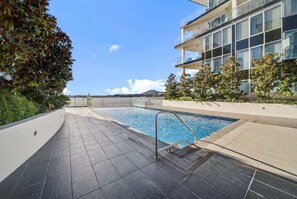 A swimming pool with a metal ladder, surrounded by a tiled deck, and adjacent to a modern building with large windows. - Spacious Lakefront Apartment with Pool Access (Kingston)