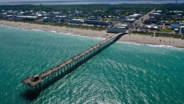 An aerial view of a coastal town with a long pier extending into the turquoise sea, surrounded by beach houses and a dense forest in the background.