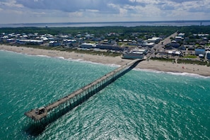 An aerial view of a coastal town with a long pier extending into the turquoise sea, surrounded by beach houses and a dense forest in the background.