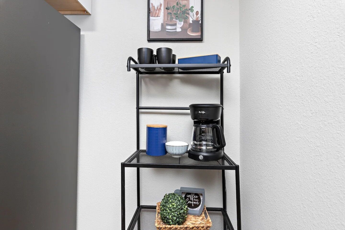 A black metal shelf unit with a coffee maker, a blue container, and a decorative plant.