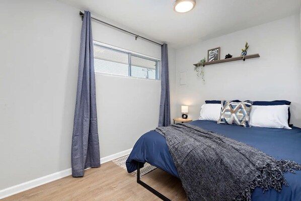 A bedroom with a bed, a bedside table with a lamp, a window with curtains, and a shelf with decorative items.