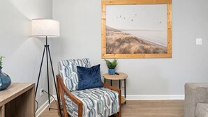 Una habitación con una silla de madera, una lámpara, una mesita con una planta y un cuadro en la pared.