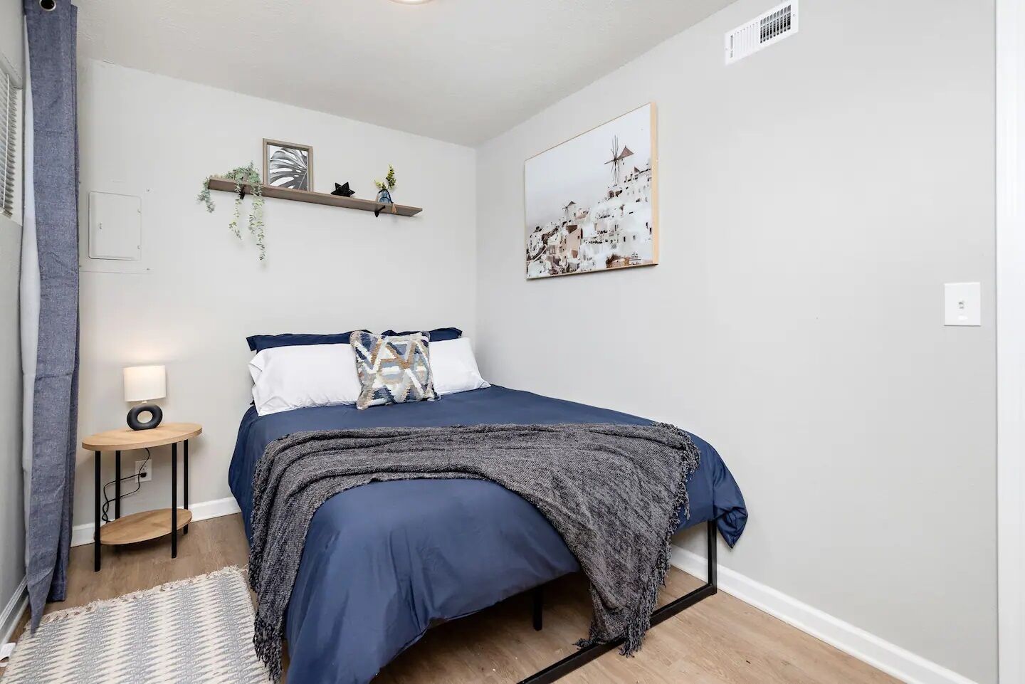 Un dormitorio con una cama, una mesita de noche con lámpara, un estante en la pared con plantas y un cuadro enmarcado.