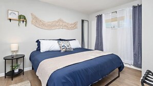 Un dormitorio con cama, mesita de noche con lámpara, espejo en la pared y ventana con cortinas.
