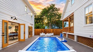 Une piscine avec des chaises longues, une terrasse en bois et une vue sur une maison dotée d’une terrasse et illuminée de guirlandes lumineuses.