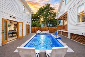 A swimming pool with lounge chairs, a wooden deck, and a view of a house with a deck and string lights. - The Grand Augustine l Heated Pool l Walk to DNTN (St. Augustine)