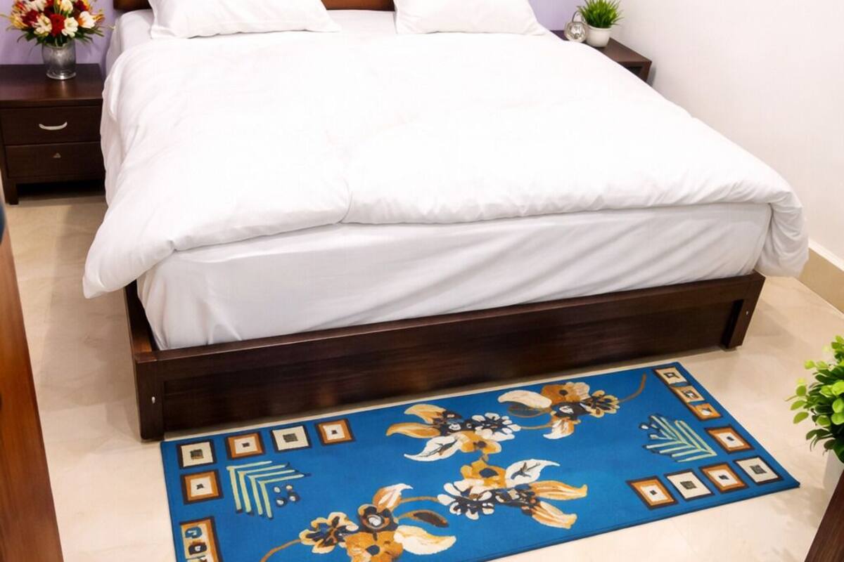 A bedroom with a wooden headboard, a bed with white bedding, a blue floral rug, and a small potted plant.