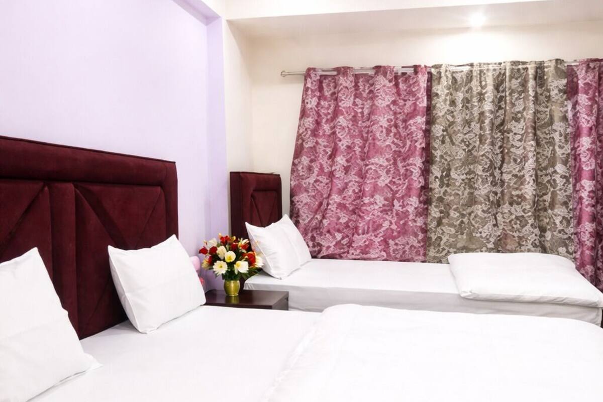 A hotel room with two beds, a nightstand with a vase of flowers, and a window with patterned curtains.