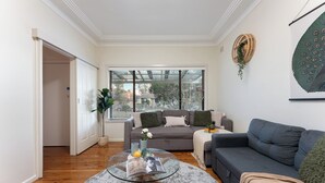 Un salon avec une table basse en verre, un canapé gris et un fauteuil gris. On y trouve une fenêtre avec des stores, une plante et un élément de décoration fixé au mur.
