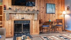A cozy living room with a wooden wall, a fireplace, a television mounted above it, and a dining area with two chairs and a table.