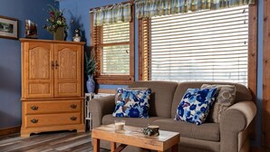 A living room with a brown sofa, two blue floral cushions, a wooden coffee table, a wooden cabinet, and a window with blinds.