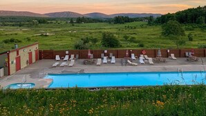 A pool area with lounge chairs and umbrellas, surrounded by a wooden fence, with a view of green fields and distant hills.