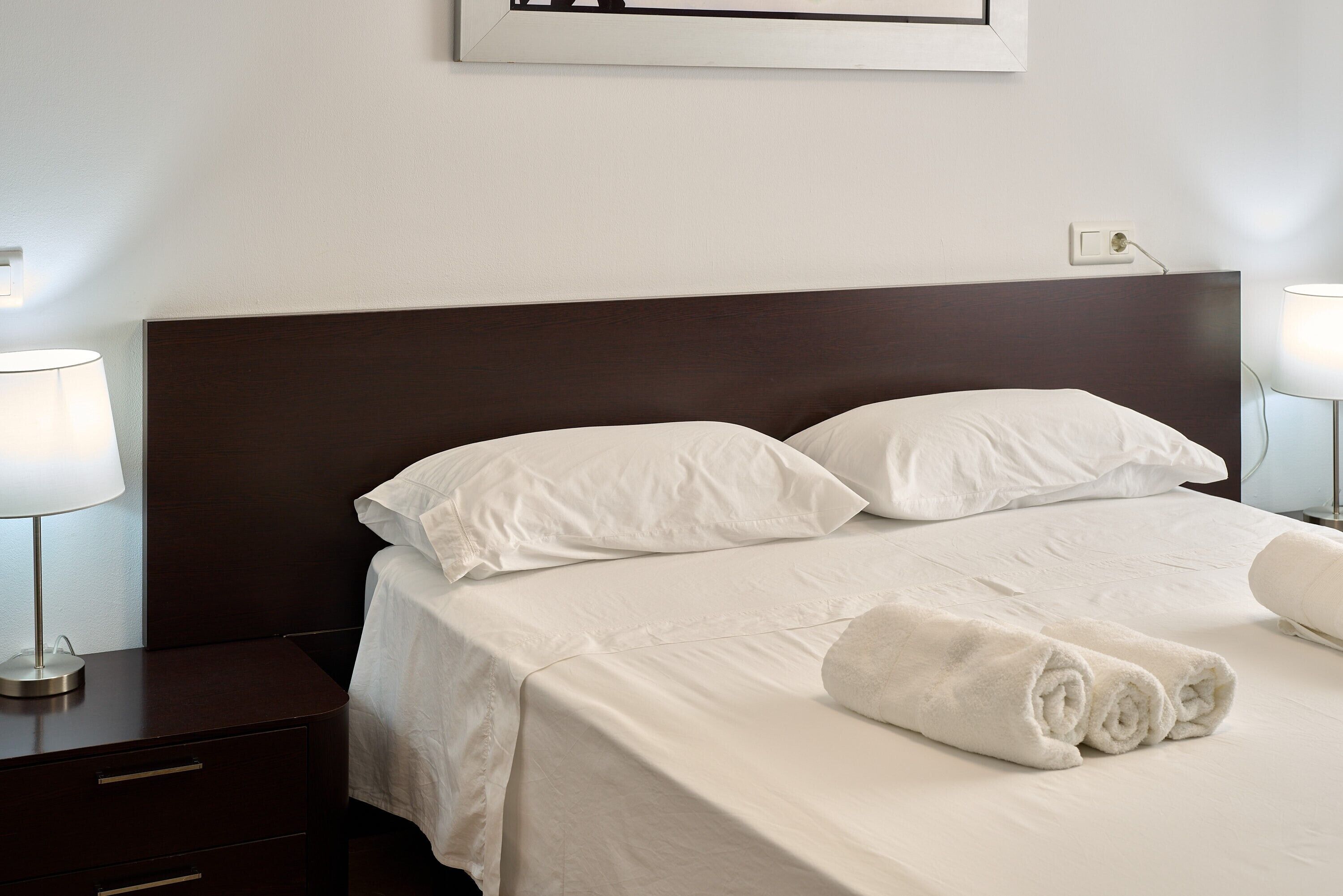 Una cama bien hecha con sábanas blancas, cabecera oscura y una lámpara de noche sobre una mesita de madera.