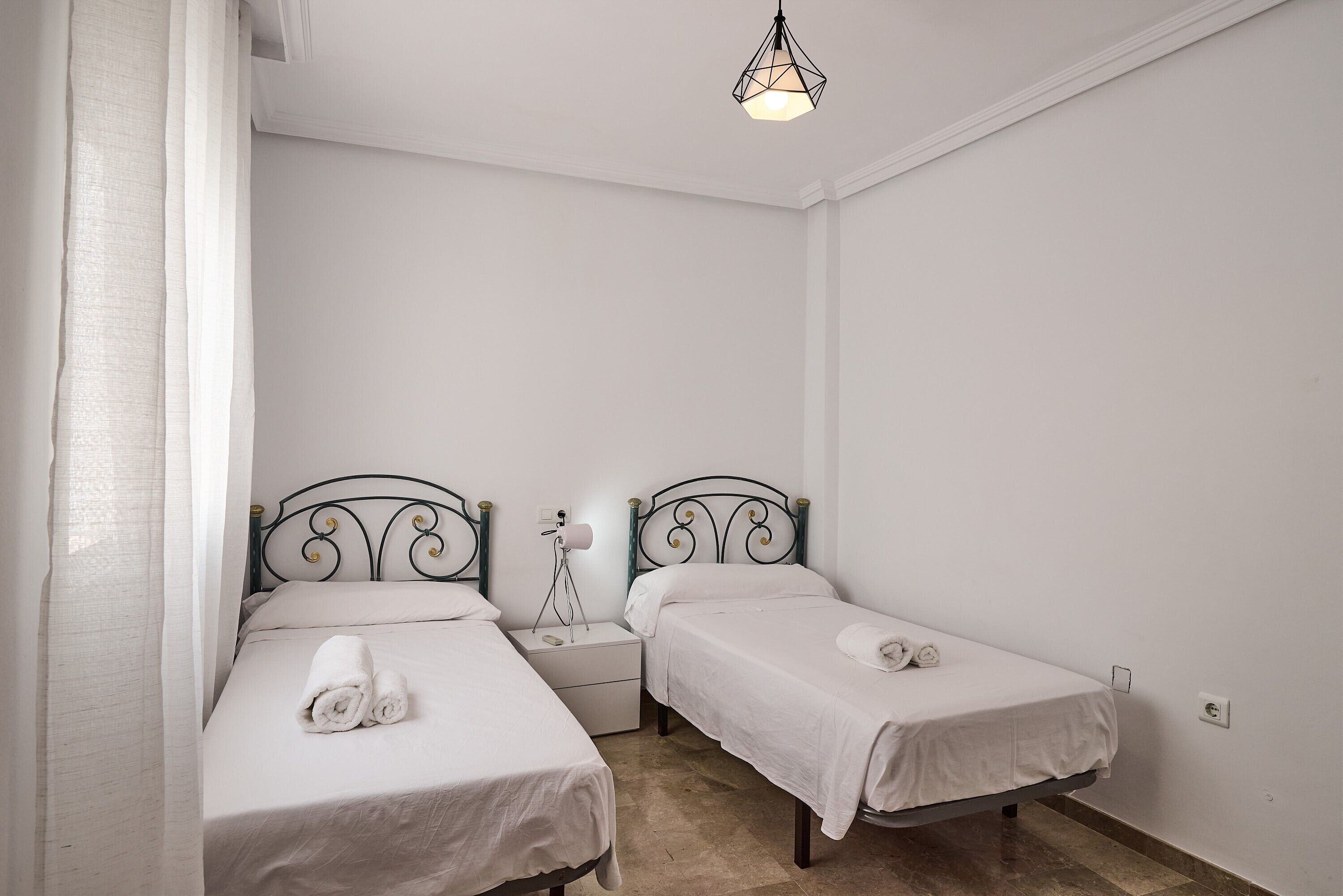 Una habitación pequeña y limpia con dos camas individuales, una mesita de noche, una lámpara y un candelabro.