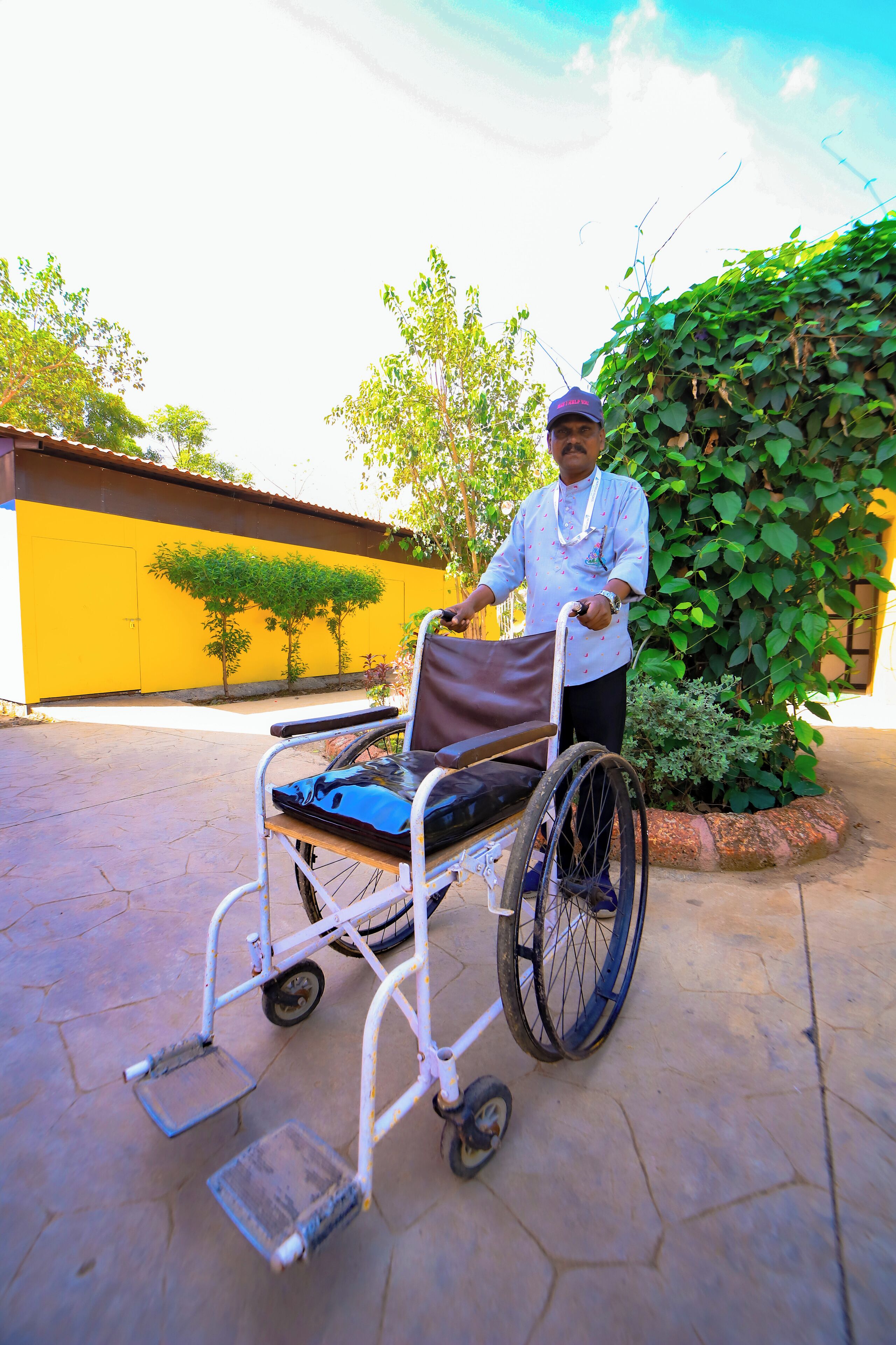 A person in a wheelchair with a tray, a paved outdoor area, and a yellow wall with trees.