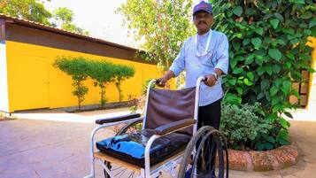 A person in a wheelchair with a tray, a paved outdoor area, and a yellow wall with trees.