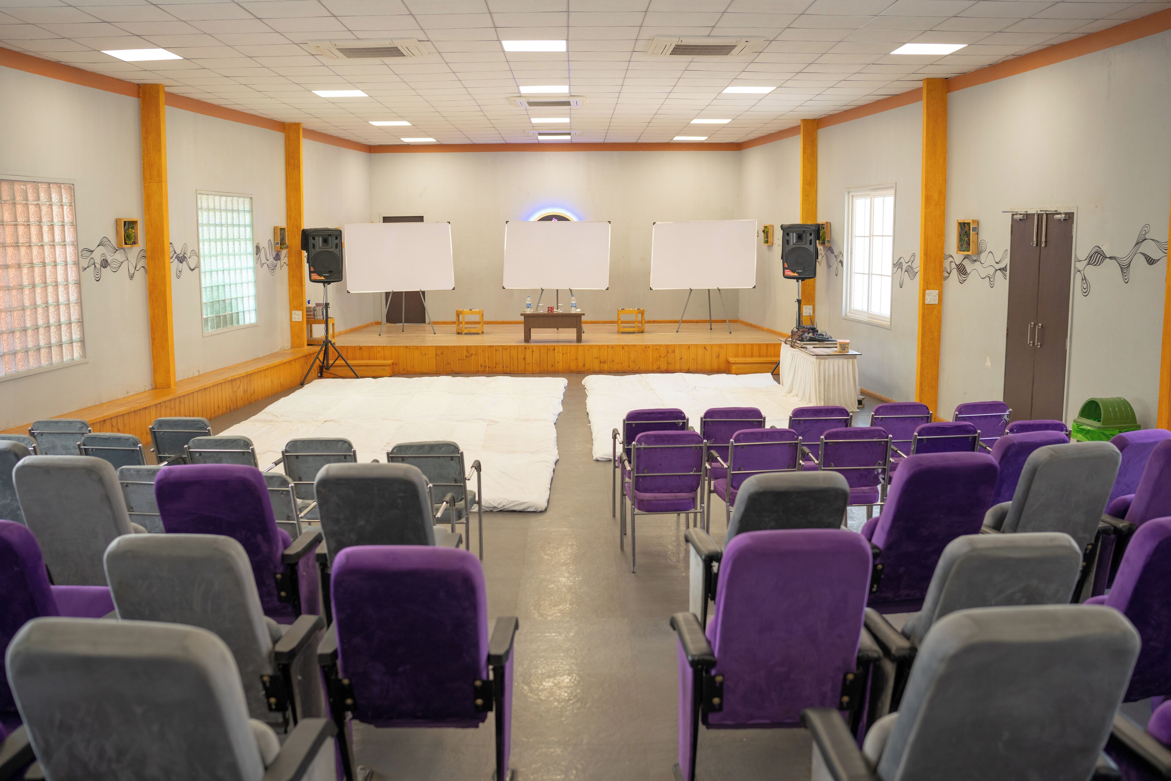Ein Konferenzraum mit einer Bühne, Whiteboards und Reihenweise violett-grauer Stühle.