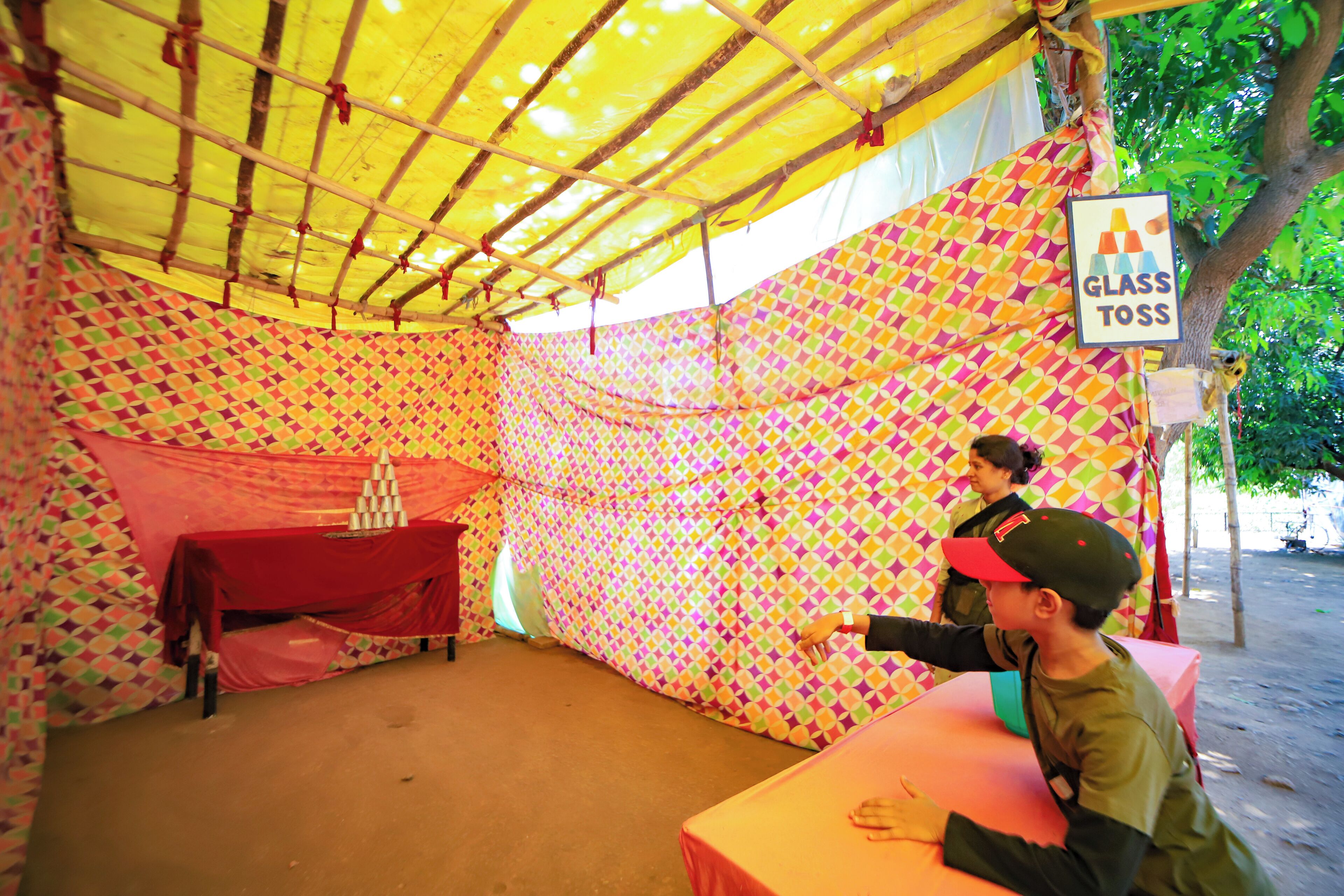 A tent with a colorful geometric pattern, a table with a red cloth, and a sign indicating a glass toss game.