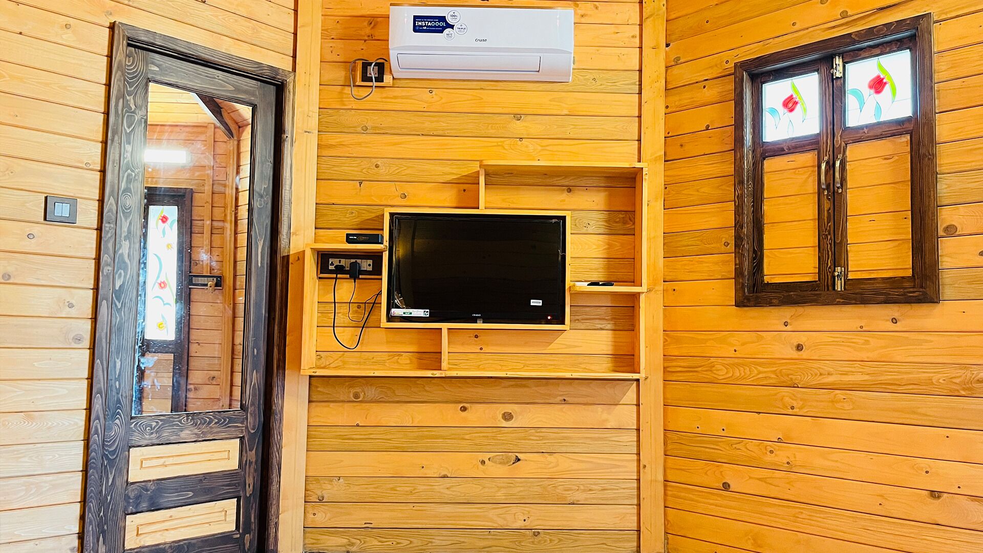 A wooden cabin interior with a television mounted on the wall, a door with a decorative glass panel, and a window with a floral design.