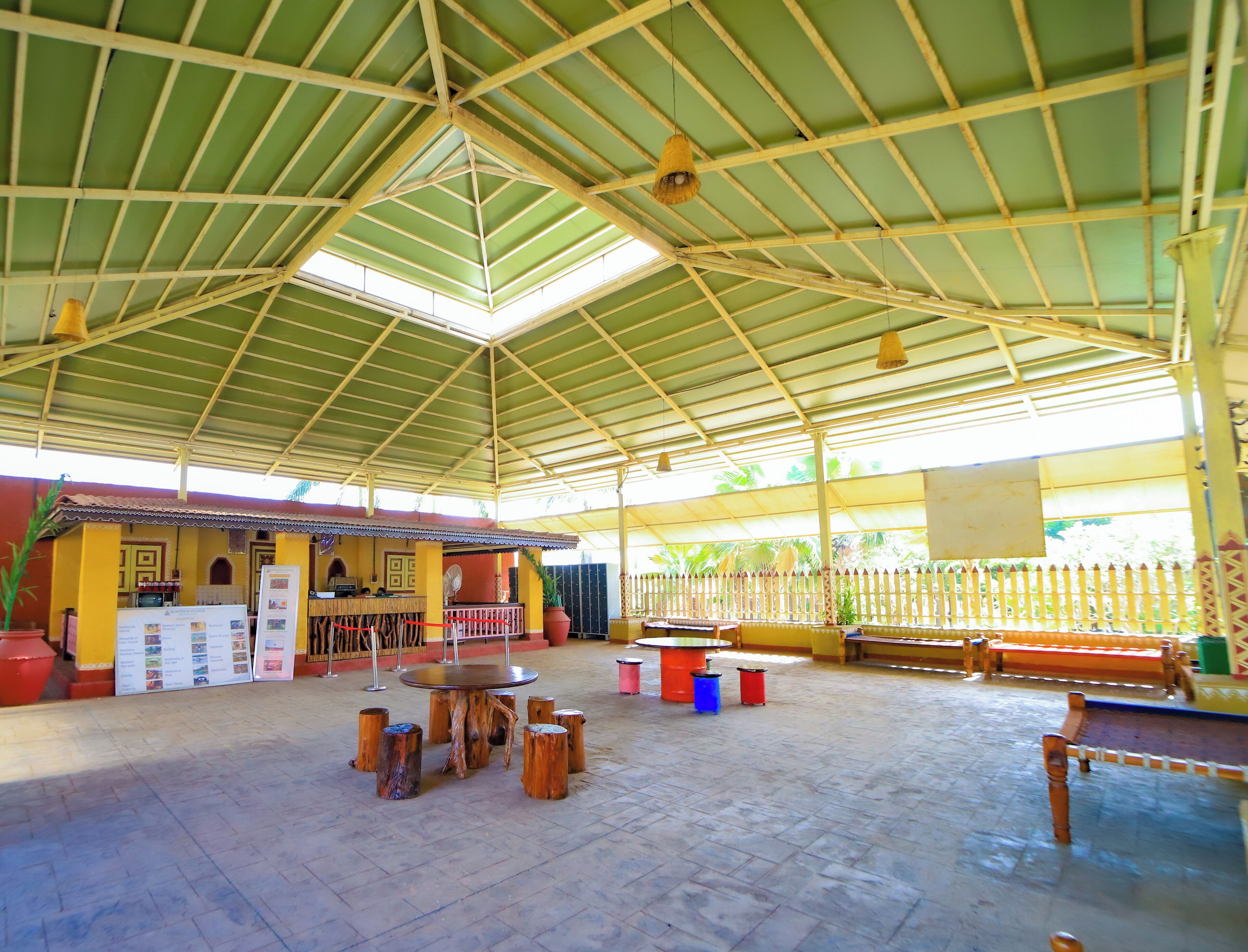 A covered outdoor area with a high ceiling, a central table surrounded by wooden stools, and a seating area with benches.