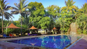 A swimming pool with lounge chairs and umbrellas, surrounded by palm trees and greenery. - Monteria Village  (Khalapur)