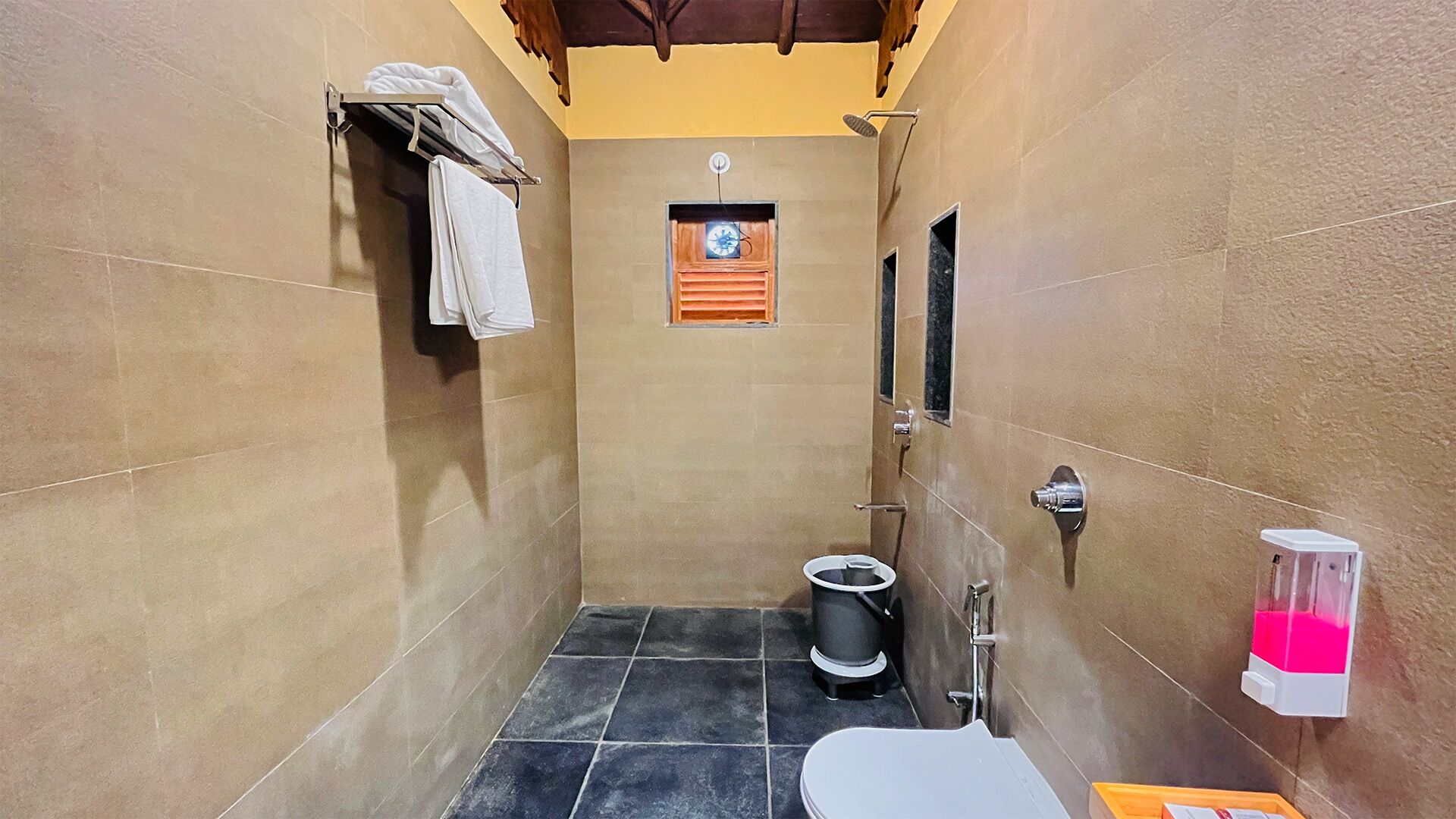 A small, narrow bathroom with a toilet, a trash bin, and a hand dryer. There is a towel rack on the wall and a small window with a clock.