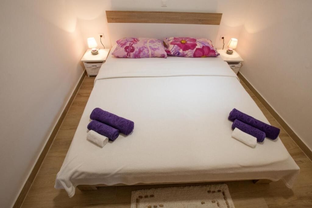 A neatly made bed with white sheets and two pillows, two purple towels, and two bedside lamps on wooden nightstands.