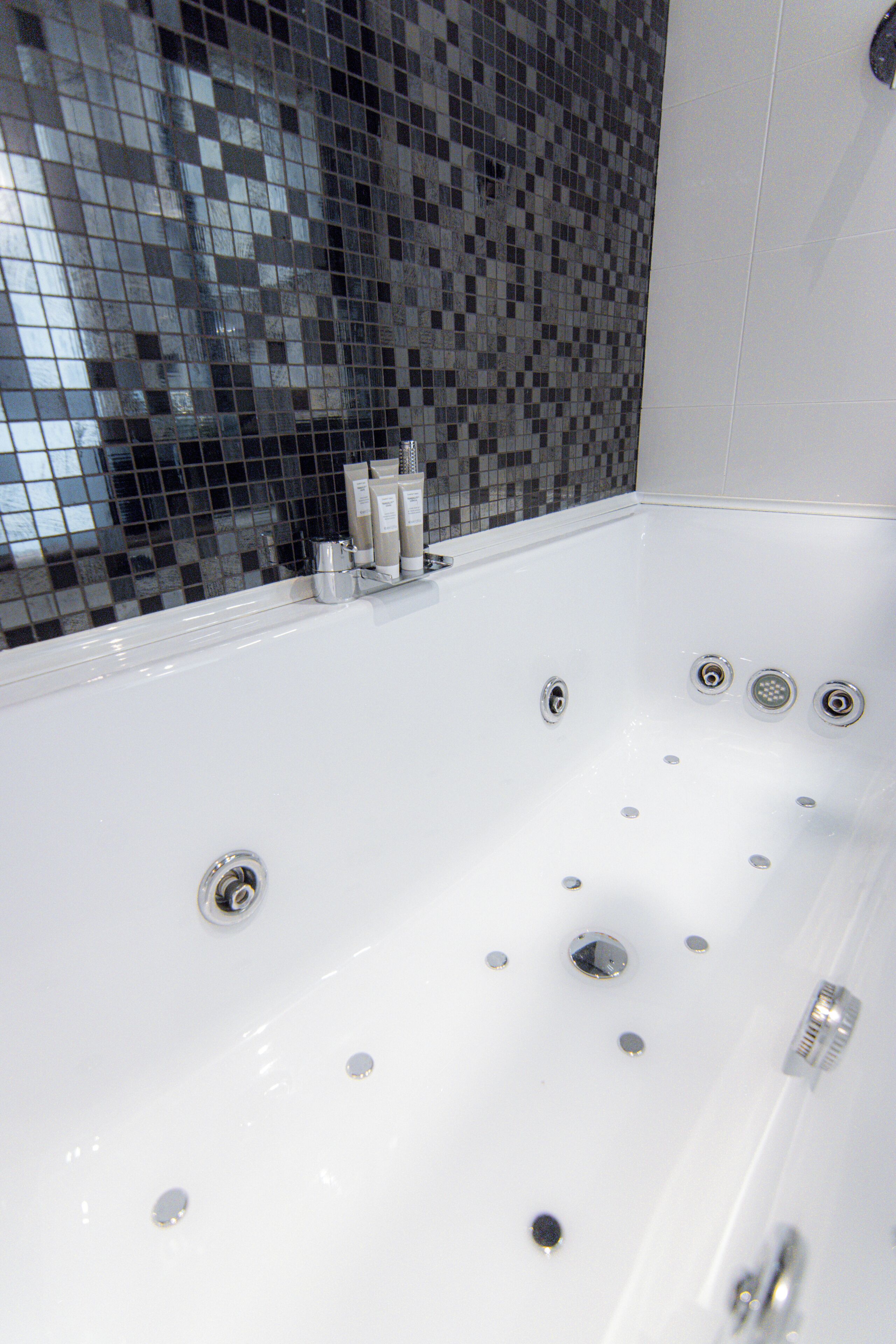 Una vasca da bagno bianca con numerosi rubinetti e una parete piastrellata con motivo a mosaico.