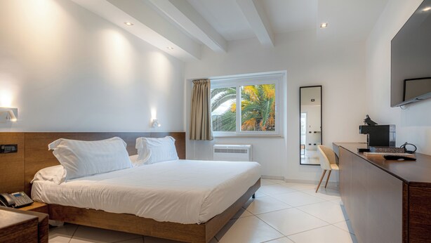 Een moderne hotelkamer met een groot bed, een bureau, een stoel, een spiegel en een raam met uitzicht op palmbomen.