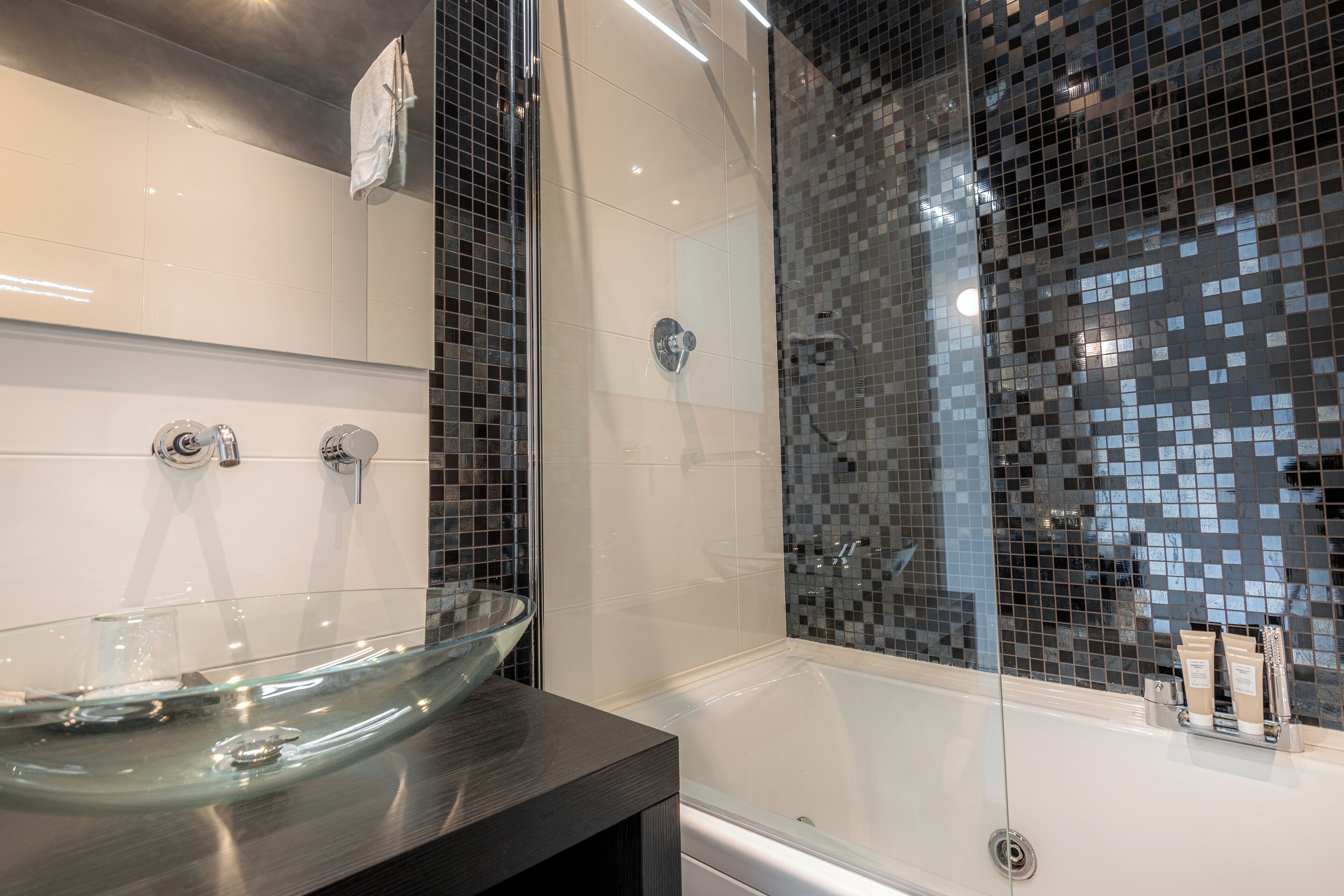 A modern bathroom with a glass vessel sink, a bathtub, and a shower area with mosaic tiles.