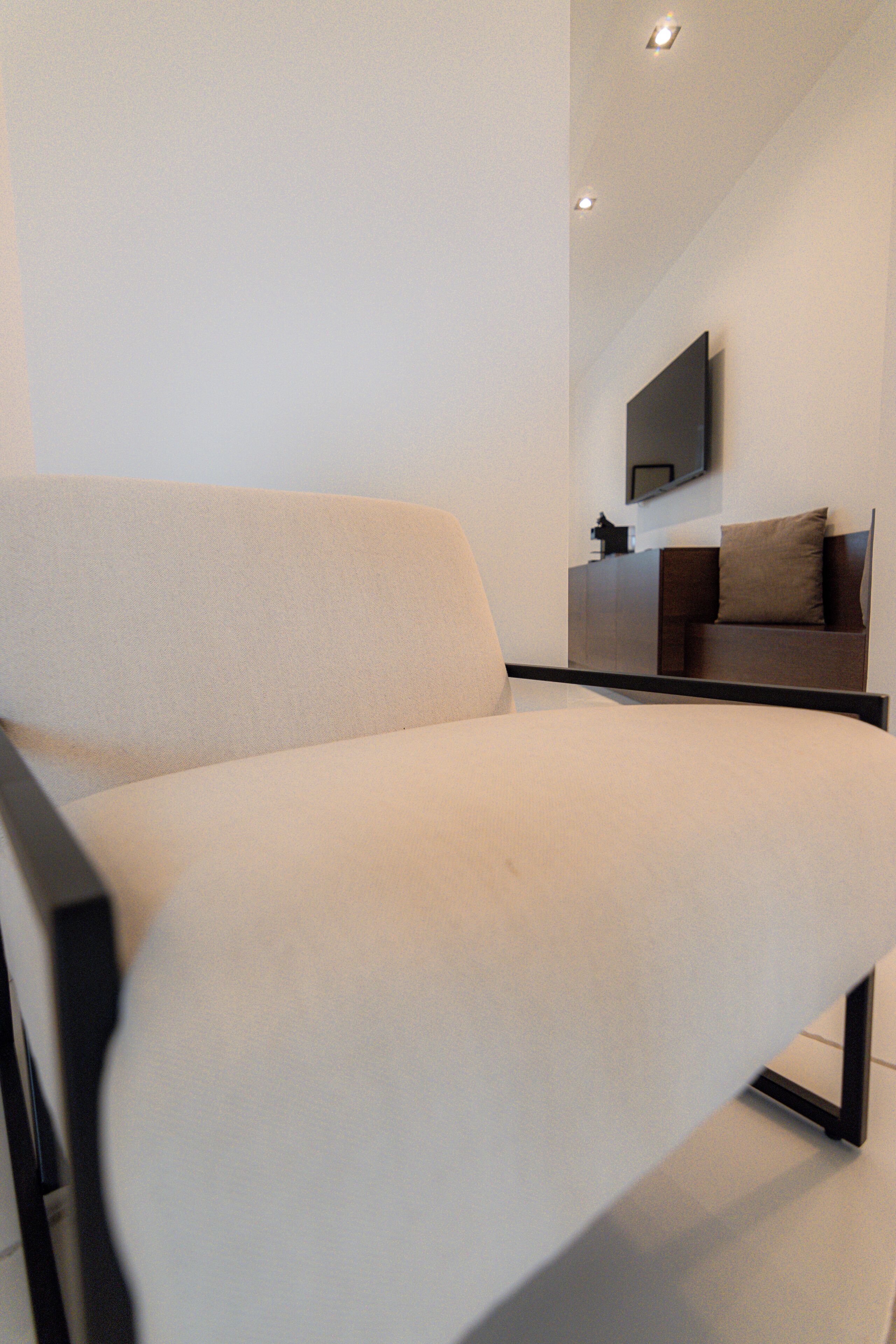 A modern, minimalist living room with a sofa, a flat-screen TV mounted on the wall, and a small table with a lamp.