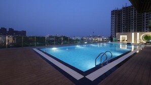 Una piscina en la azotea con terraza de madera, rodeada por barandilla de vidrio y vistas al horizonte urbano al atardecer.