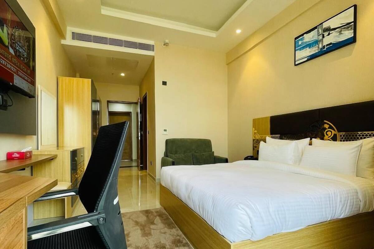 A hotel room with a bed, a desk with a chair, a TV, and a painting on the wall.