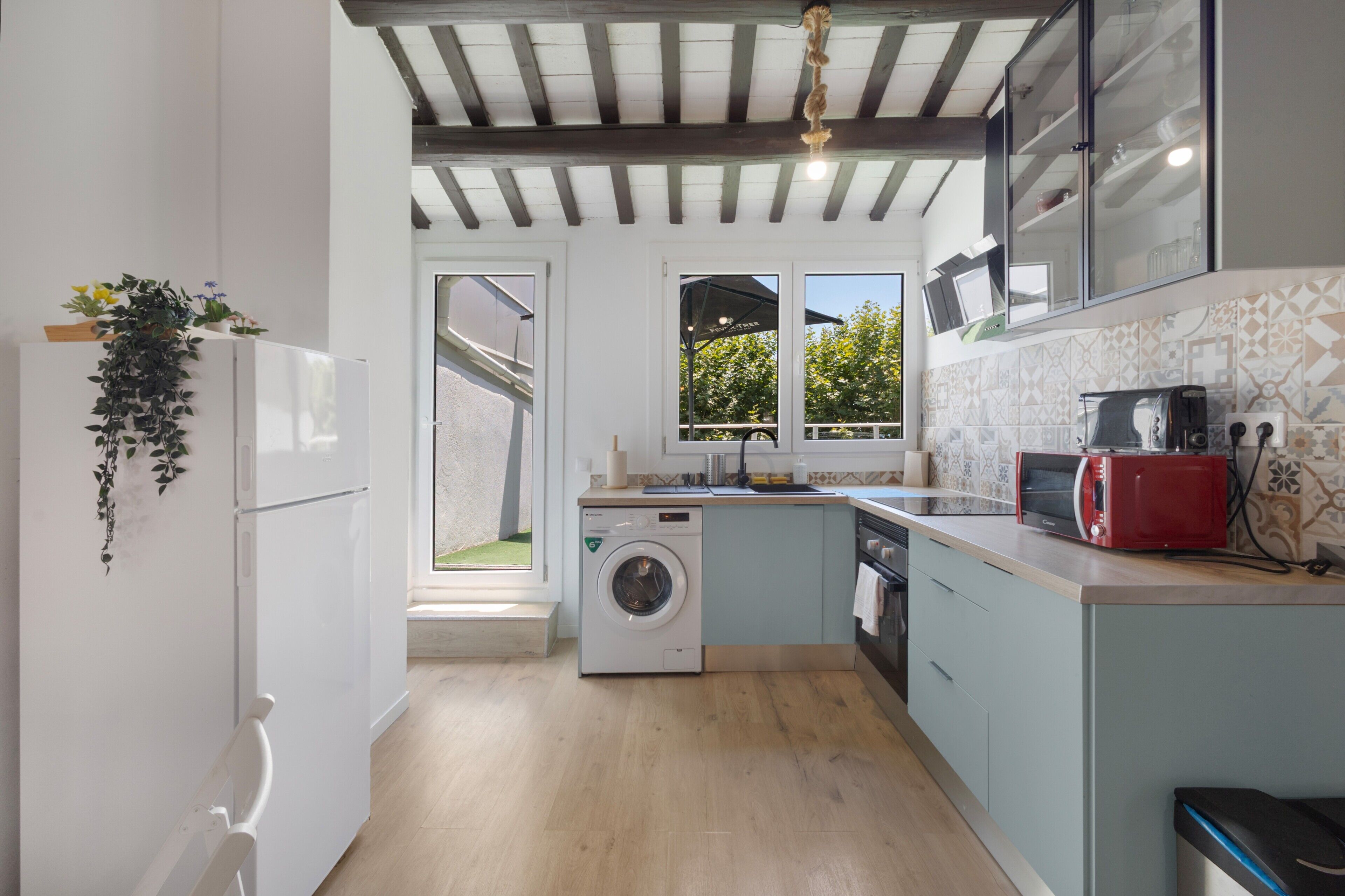 Une cuisine compacte équipée d’un lave-linge, d’un grille-pain rouge et d’une fenêtre donnant sur un jardin.