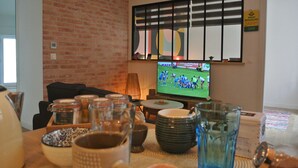 A living room with a brick wall, a television displaying a sports game, and various kitchen items on the table.