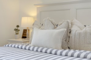 Una cama bien hecha con ropa de cama a rayas, un cabecero blanco y una mesita de noche con una lámpara y una planta pequeña.